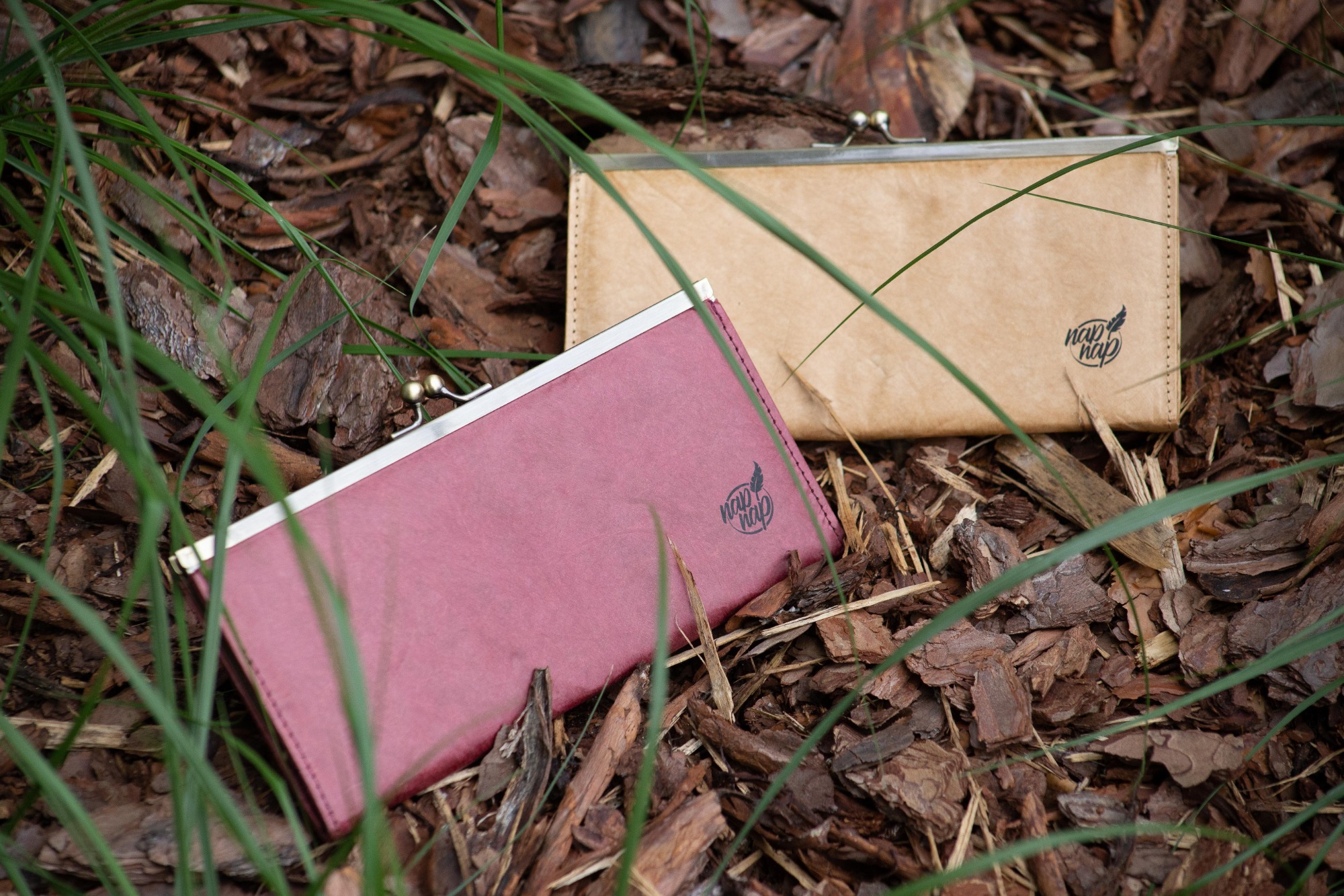 Dwa damskie portfele na zatrzask, jeden bordowy, drugi beżowy, leżą na ściółce z kory drzewnej, z fragmentami zielonej trawy w kadrze.