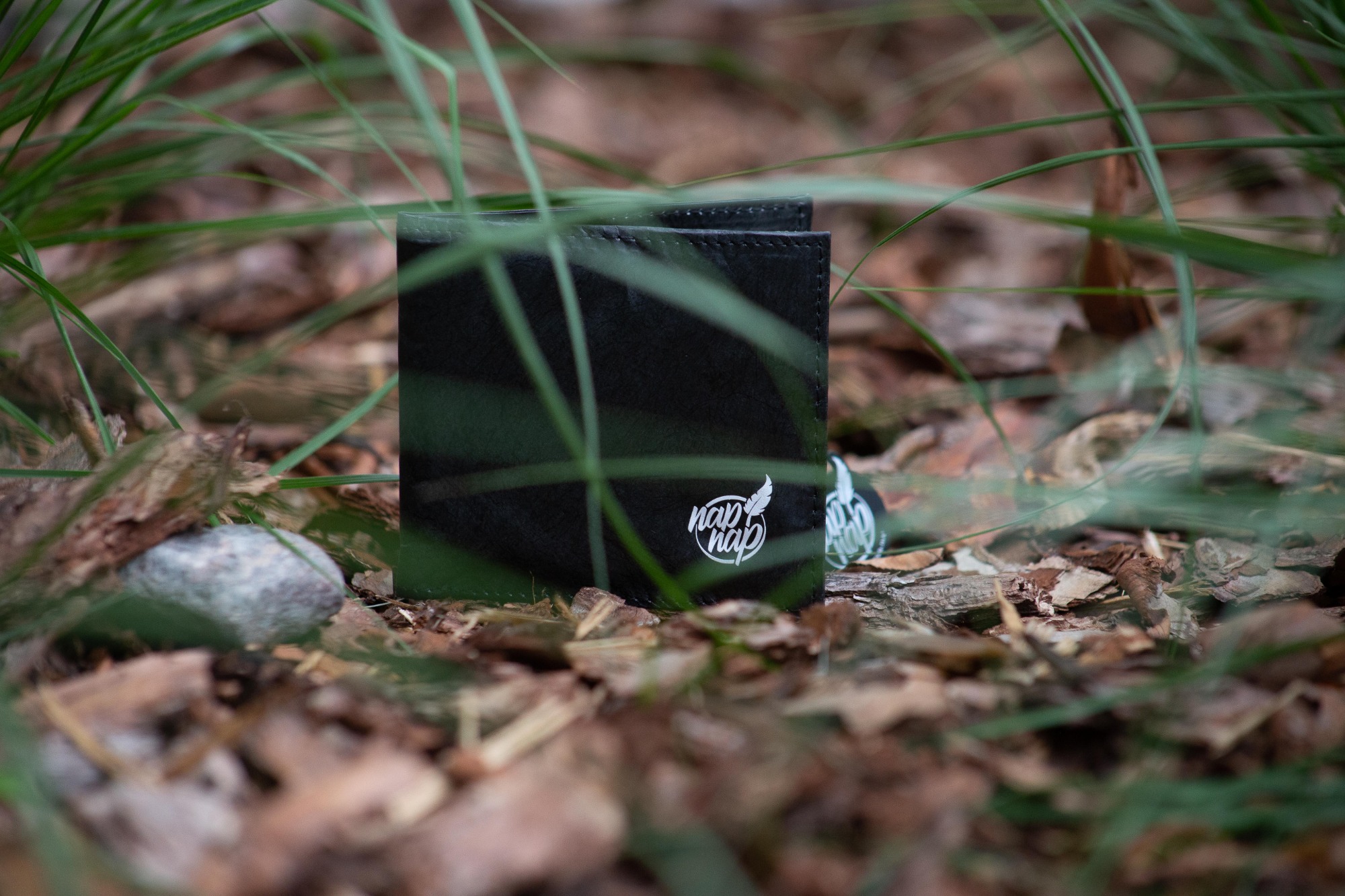 Czarny portfel z logo firmy na tle ściółki leśnej i źdźbeł trawy.