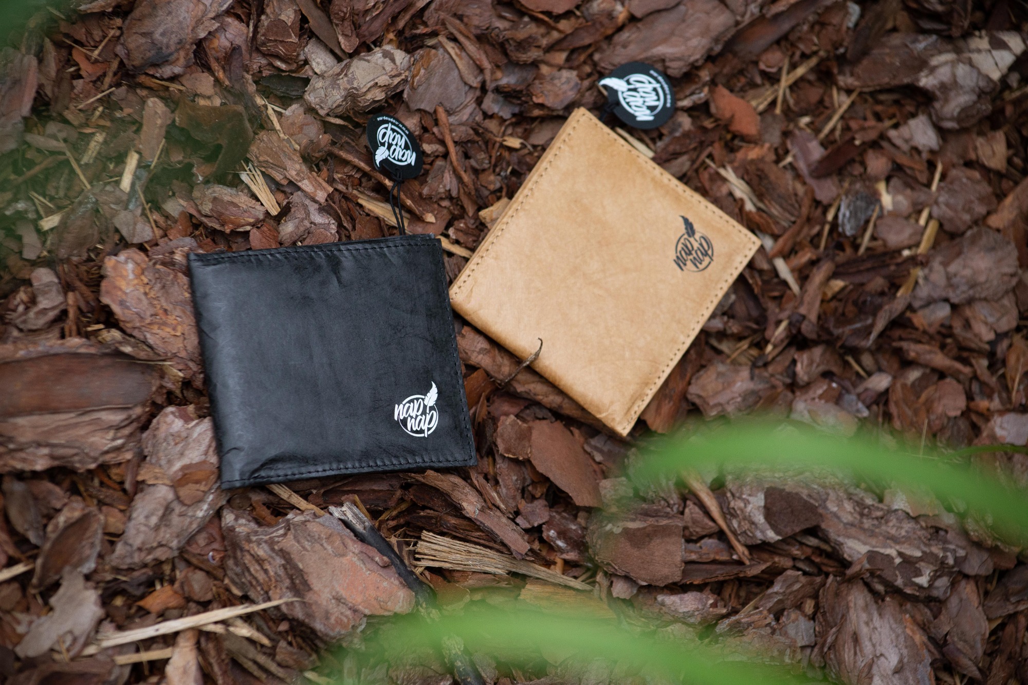 Dwa portfele, czarny i beżowy, leżące na ściółce z kory drzewnej, z widocznymi metkami z logo firmy.