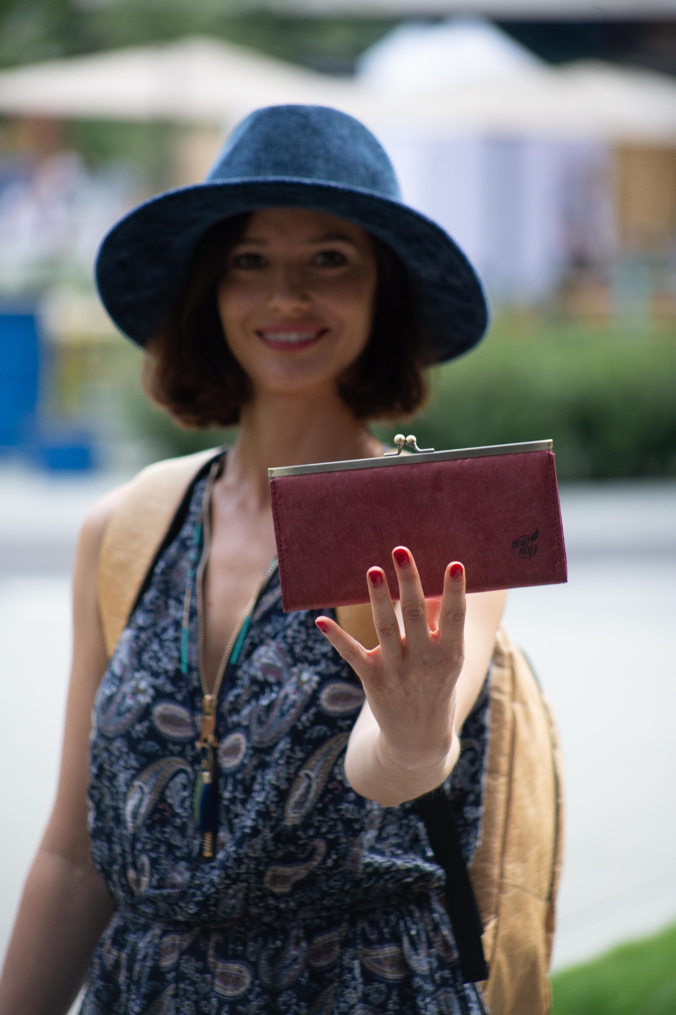 Uśmiechnięta kobieta w niebieskim kapeluszu prezentuje bordowy portfel na tle rozmytej zieleni, z widocznym logo producenta na portfelu.