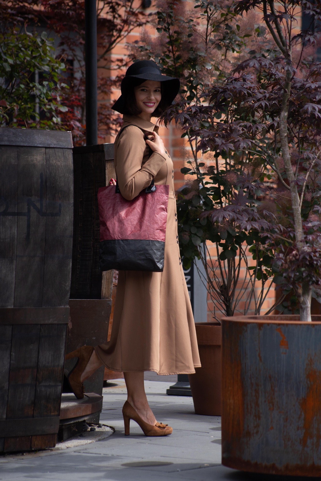 Uśmiechnięta kobieta w brązowej sukience, czarnym kapeluszu i brązowych butach na obcasie trzyma czerwoną torbę na tle roślinności i drewnianych beczek.