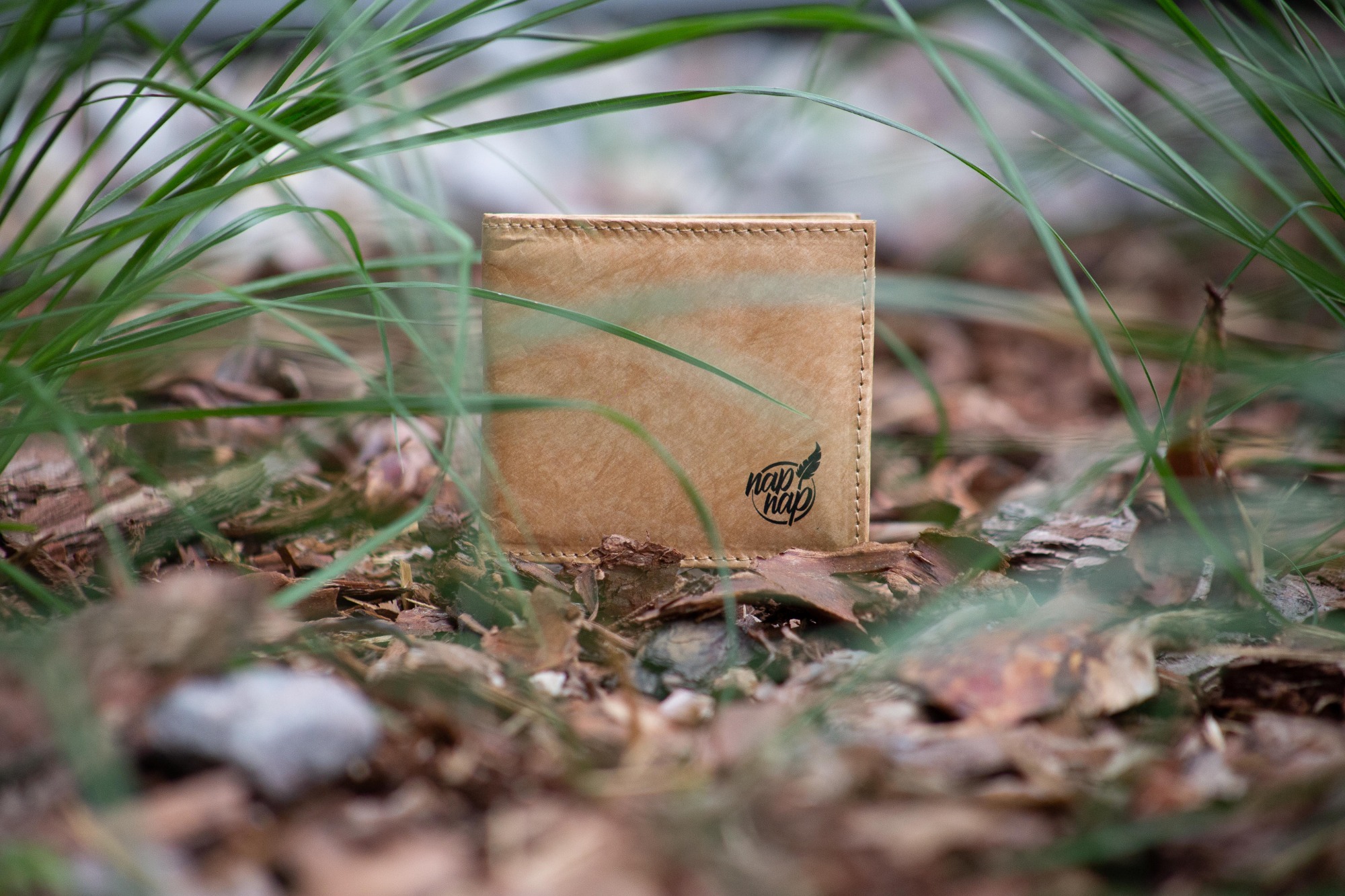 Portfel z ekologicznego materiału, z logo 'nap nap', umieszczony na ściółce leśnej, otoczony źdźbłami trawy.