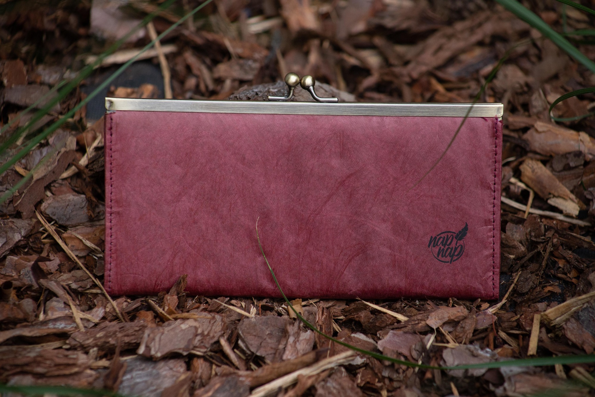 Burgundowy portfel damski z metalowym zapięciem, leżący na ściółce z kory drzewnej, z widocznym logo 'nap nap' w lewym dolnym rogu.