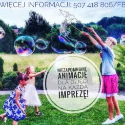 Animator w różowej spódnicy puszcza duże bańki mydlane dla dwóch dziewczynek na trawie; w tle zieleń i numer telefonu kontaktowego.