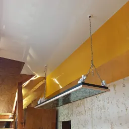 Biały, błyszczący sufit napinany z zamontowaną designerską lampą wiszącą na łańcuchach, żółta ściana w tle, fragment drewnianych schodów i boazerii.