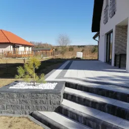 Nowoczesny taras z jasnoszarej kostki brukowej z ciemnymi obrzeżami, schody z ciemnymi krawężnikami, donica z białymi kamieniami i małą sosną na tle domu z białymi ścianami i czerwonym dachem...