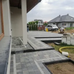 Nowo ułożony taras z kostki brukowej w odcieniach szarości, obrzeża z ciemnej kostki, widoczny fragment domu z kolumną i okno, w tle zaparkowany bus i betoniarka, teren jeszcze niezagospodarowany.