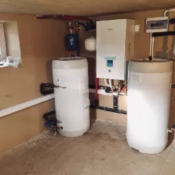 Instalacja pompy ciepła Panasonic Aquarea z dwoma zbiornikami akumulacyjnymi w piwnicy, widoczne rury i armatura, biała obudowa jednostki wewnętrznej i zbiorników na tle beżowych ścian.