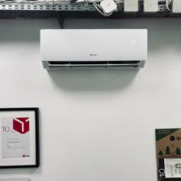 Biały klimatyzator marki Gree zamontowany na ścianie biura, widoczny wyświetlacz temperatury, pod sufitem instalacja elektryczna, obok wisi kalendarz DPD i oprawiony dyplom.