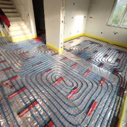 Niedokończona instalacja ogrzewania podłogowego z niebieskich rur na folii z siatką, widoczne fragmenty ścian i schodów w surowym stanie.