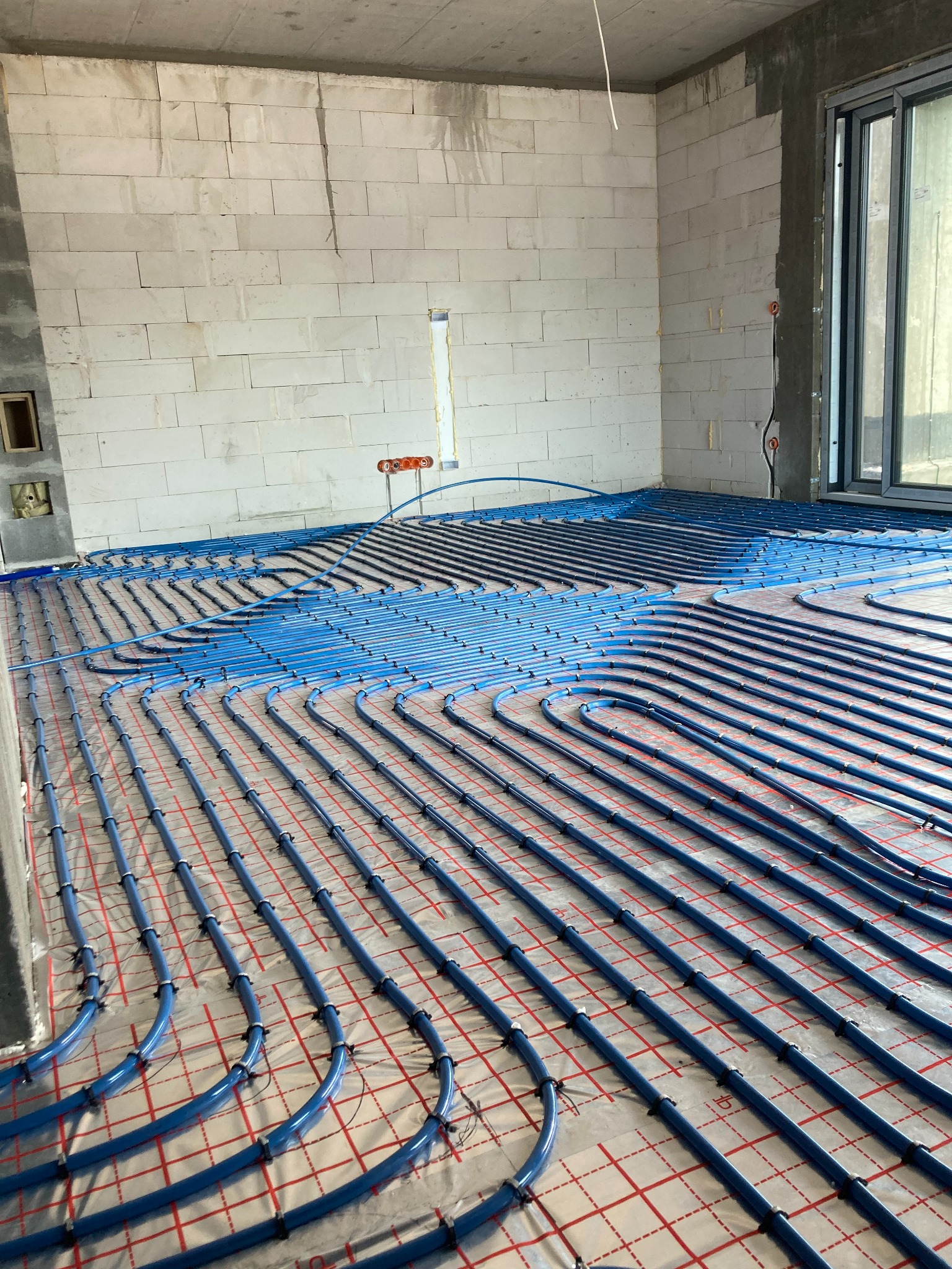 Niebieskie rury ogrzewania podłogowego rozłożone na folii z czerwoną siatką w surowym pomieszczeniu z betonowym sufitem i ścianami z białych bloczków, widoczne przyłącza.