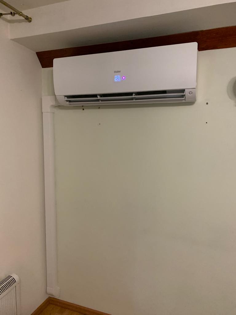 Biały klimatyzator Haier zamontowany na ścianie w pomieszczeniu, widoczny wyświetlacz z temperaturą 25 stopni Celsjusza, biała listwa maskująca przewody.