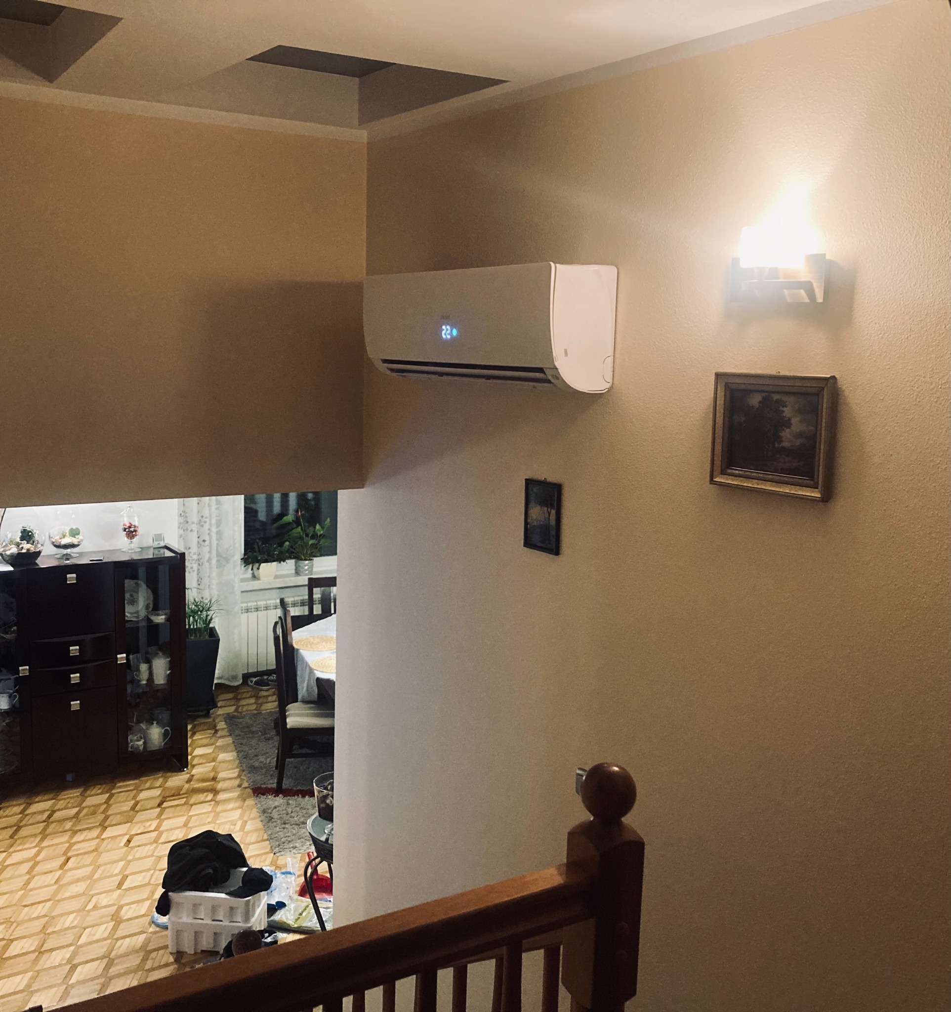 Biały klimatyzator ścienny z wyświetlaczem temperatury 22 stopnie Celsjusza, zamontowany w korytarzu nad schodami w domu z beżowymi ścianami i drewnianą balustradą.