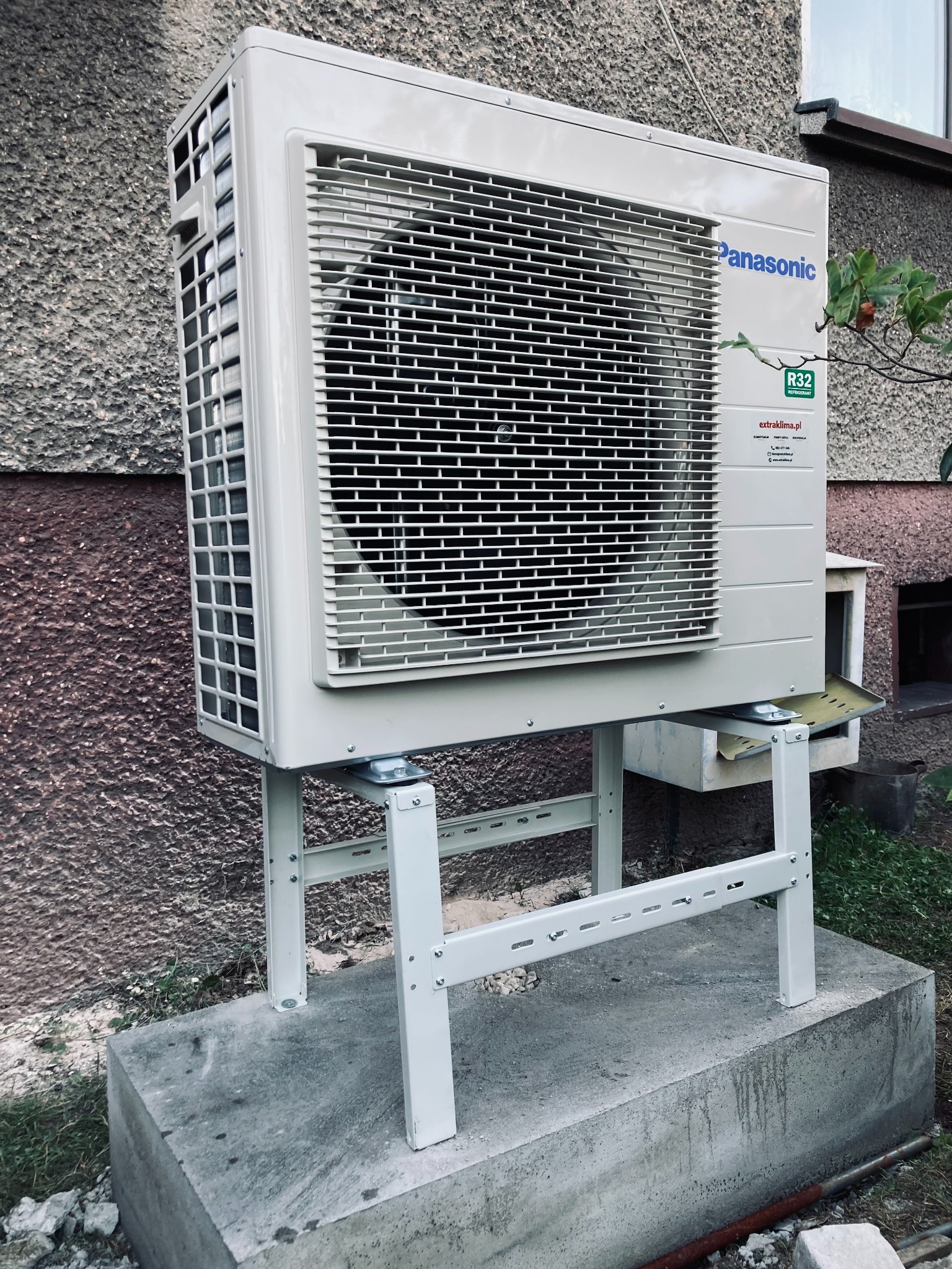 Zewnętrzna jednostka klimatyzatora Panasonic na białym, metalowym stelażu, zamontowana na betonowym fundamencie przy ścianie budynku z widocznym oknem i fragmentem roślinności.