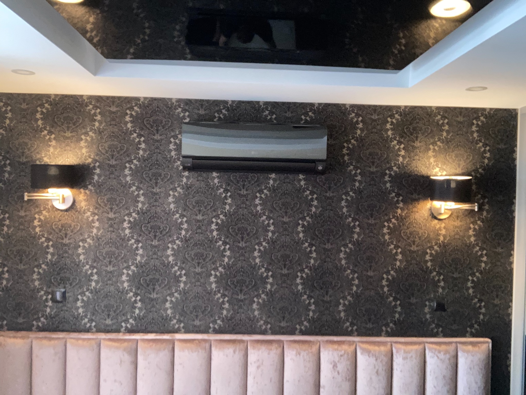 Czarny klimatyzator zamontowany na wzorzystej ścianie w Rybniku, oświetlony dwoma czarnymi kinkietami, widoczny fragment wezgłowia łóżka.