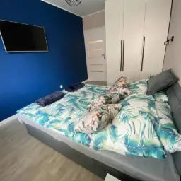 Sypialnia z łóżkiem w pościeli w tropikalne wzory, na niebieskiej ścianie wisi telewizor, a obok łóżka stoi biała szafa.