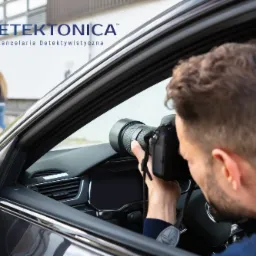 Mężczyzna z aparatem fotograficznym z dużym obiektywem robi zdjęcia z wnętrza samochodu, w tle para spacerująca po chodniku i logo firmy.