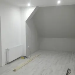 Pomieszczenie na poddaszu po malowaniu ścian na biało i szaro, z nową podłogą imitującą drewno, grzejnikiem pod oknem i listwami przypodłogowymi przygotowanymi do montażu.