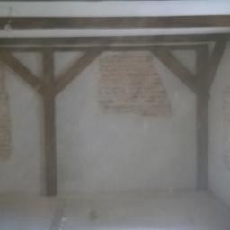 uslugi ogolnobudowlane MATEK - Surowe wnętrze poddasza z odsłoniętymi fragmentami cegieł na ścianach, drewnianą konstrukcją nośną i grzejnikiem pod oknem, w trakcie przygotowania do prac wykończeniowych.