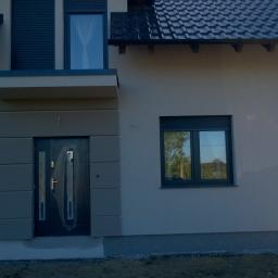 uslugi ogolnobudowlane MATEK - Elewacja domu jednorodzinnego w Sulmierzycach z szarą stolarką okienną i drzwiami wejściowymi z dekoracyjnymi panelami, widoczne fragmenty dachu i rynny.