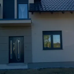 Elewacja domu jednorodzinnego w Sulmierzycach z szarą stolarką okienną i drzwiami wejściowymi z dekoracyjnymi panelami, widoczne fragmenty dachu i rynny.