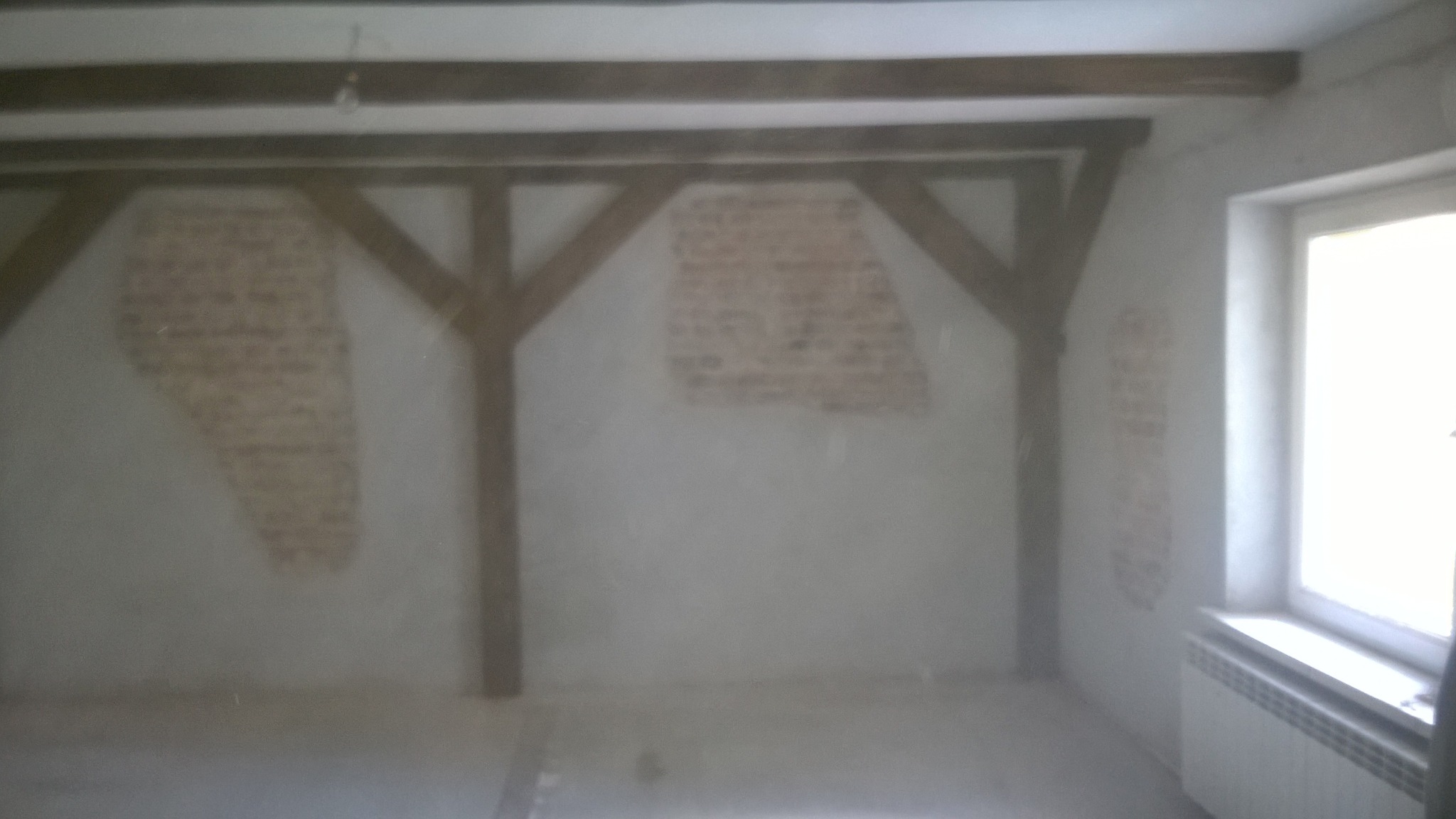 Surowe wnętrze poddasza z odsłoniętymi fragmentami cegieł na ścianach, drewnianą konstrukcją nośną i grzejnikiem pod oknem, w trakcie przygotowania do prac wykończeniowych.