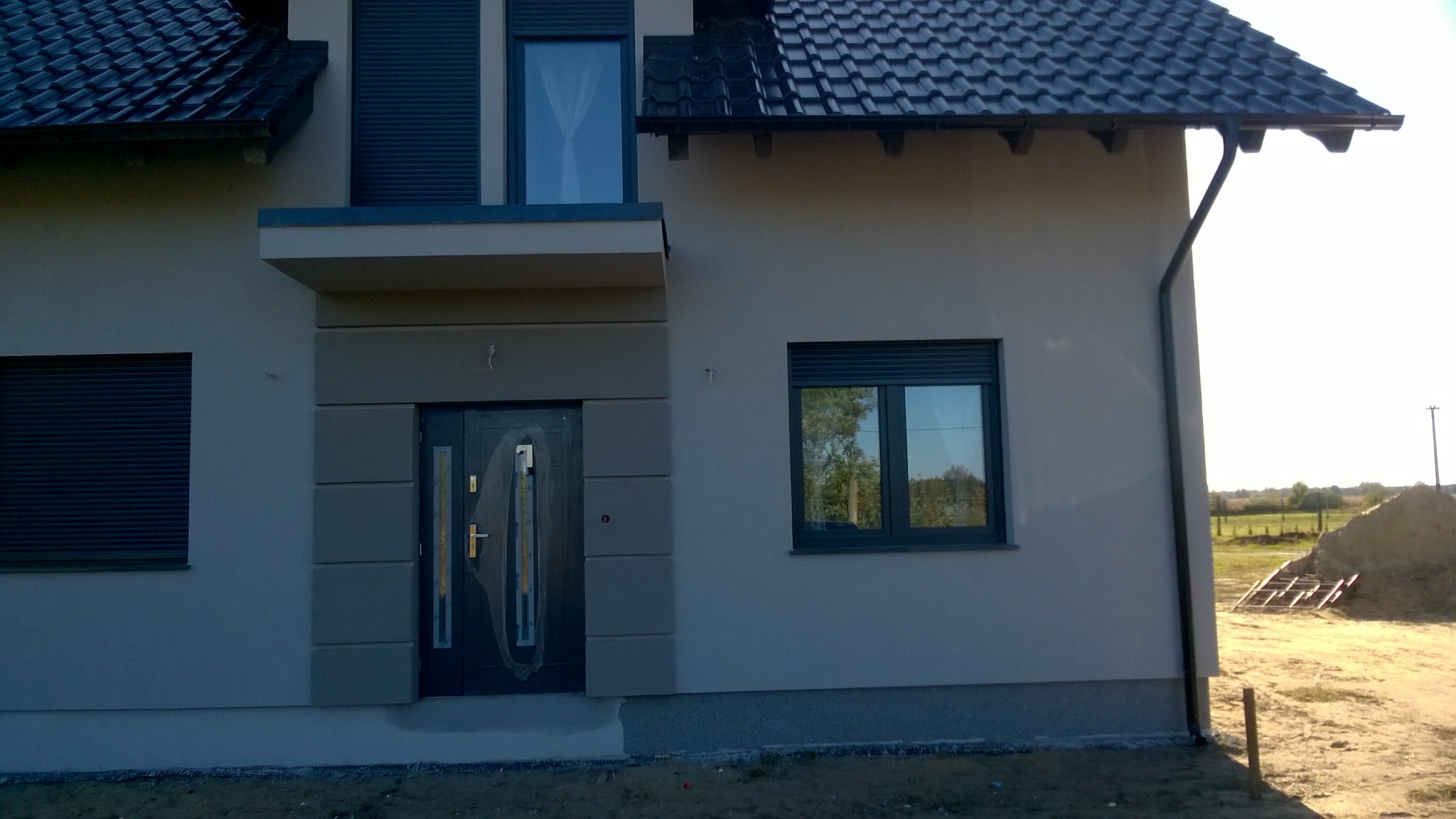 Elewacja domu jednorodzinnego w Sulmierzycach z szarą stolarką okienną i drzwiami wejściowymi z dekoracyjnymi panelami, widoczne fragmenty dachu i rynny.