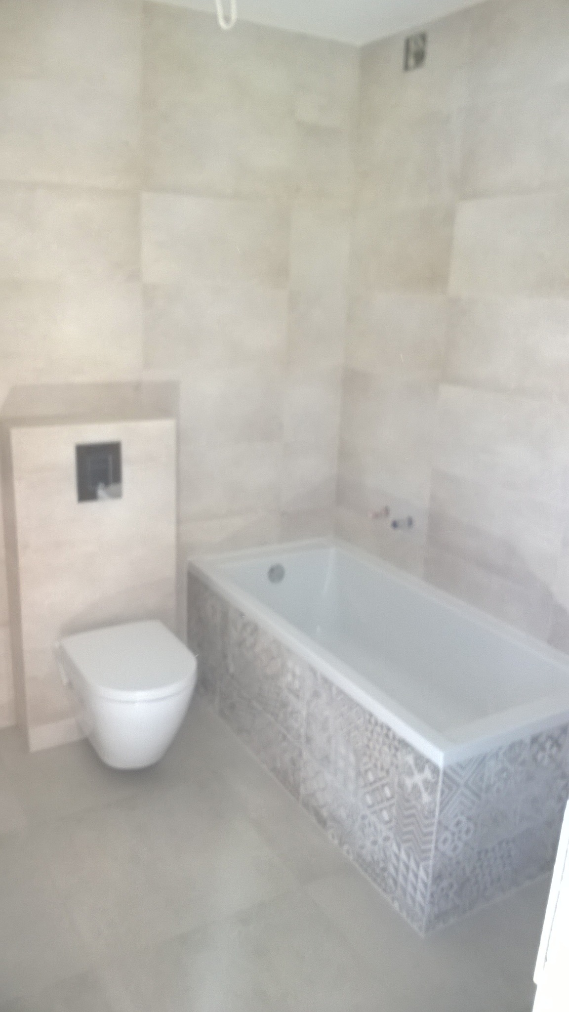 Jasna łazienka po remoncie: biała wanna z dekoracyjną obudową, podwieszana toaleta i płytki imitujące kamień na ścianach i podłodze.