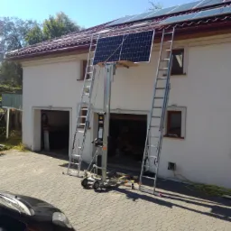 Montaż paneli słonecznych na dachu domu jednorodzinnego, widoczny podnośnik, drabiny opierają się o ścianę budynku.