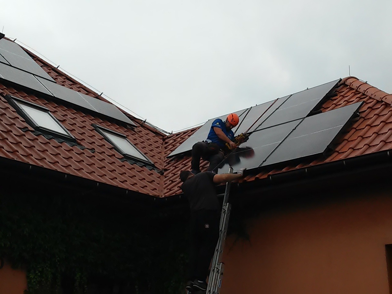 Montaż paneli fotowoltaicznych na dachu z dachówką ceramiczną; dwaj instalatorzy pracują na wysokości, jeden wierci panel, drugi asekuruje z drabiny.