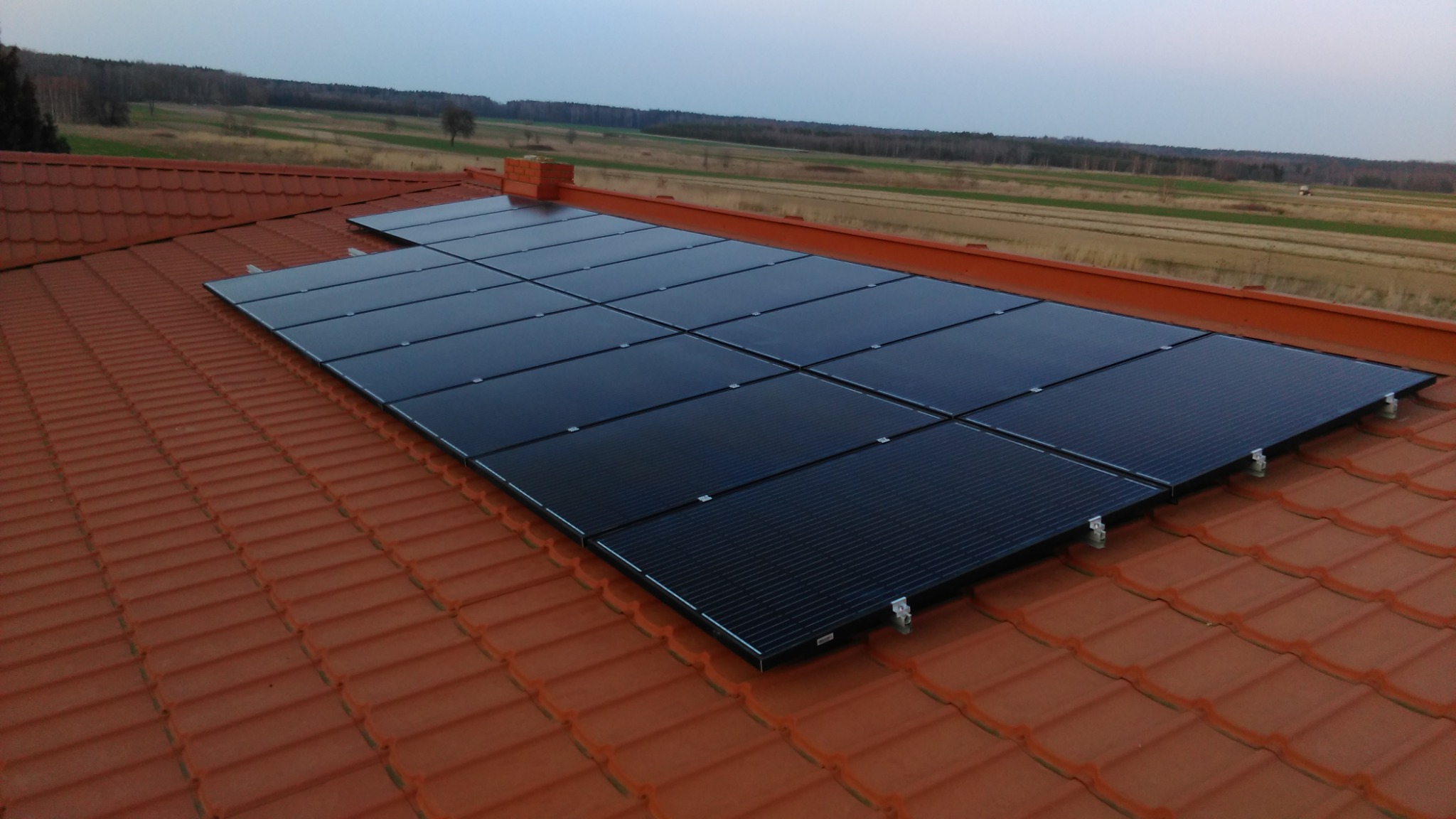 Panele słoneczne zamontowane na dachu z czerwonej dachówki, widok na pola i las w tle, pochmurne niebo.