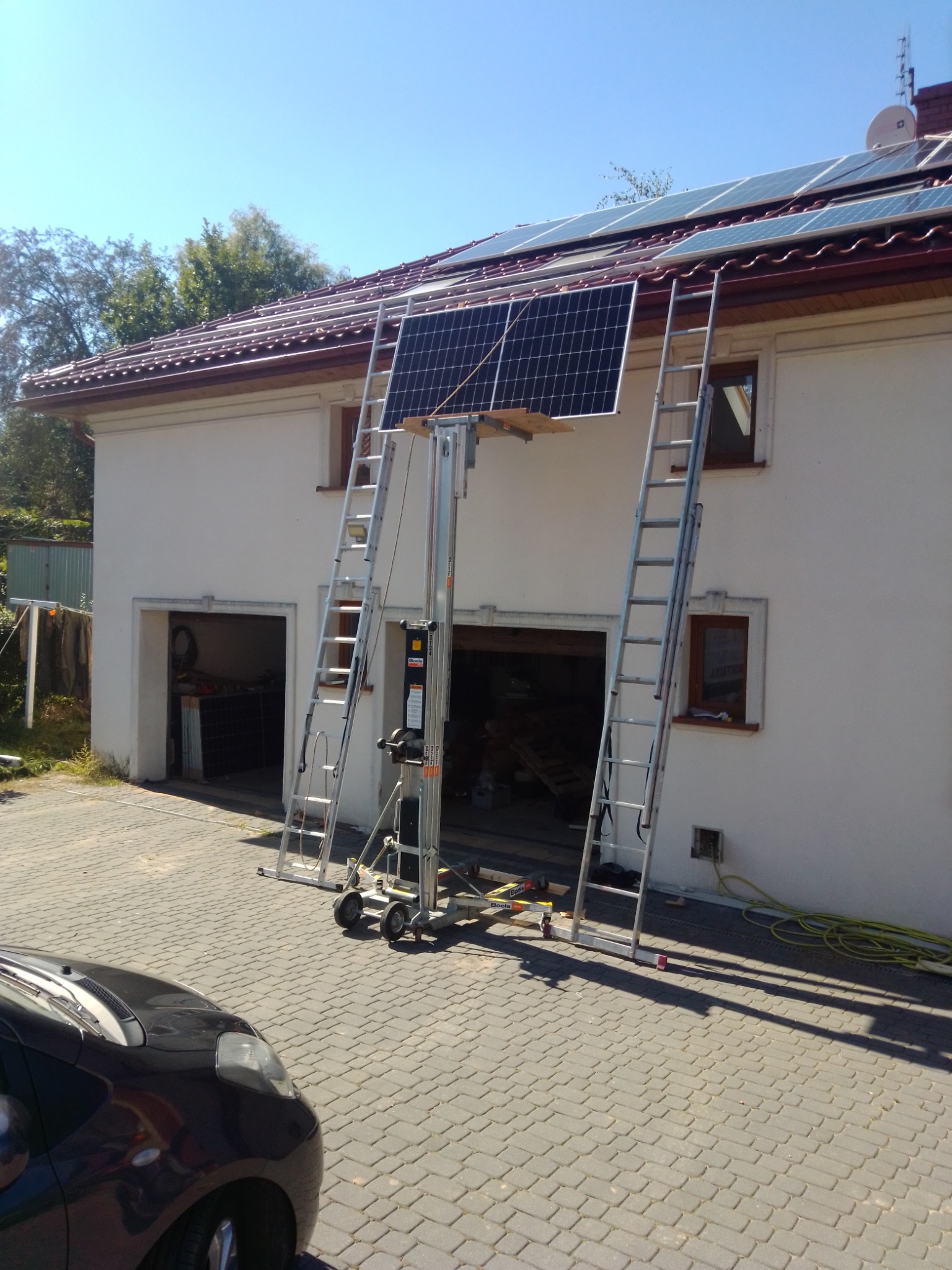 Montaż paneli słonecznych na dachu domu jednorodzinnego, widoczny podnośnik, drabiny opierają się o ścianę budynku.