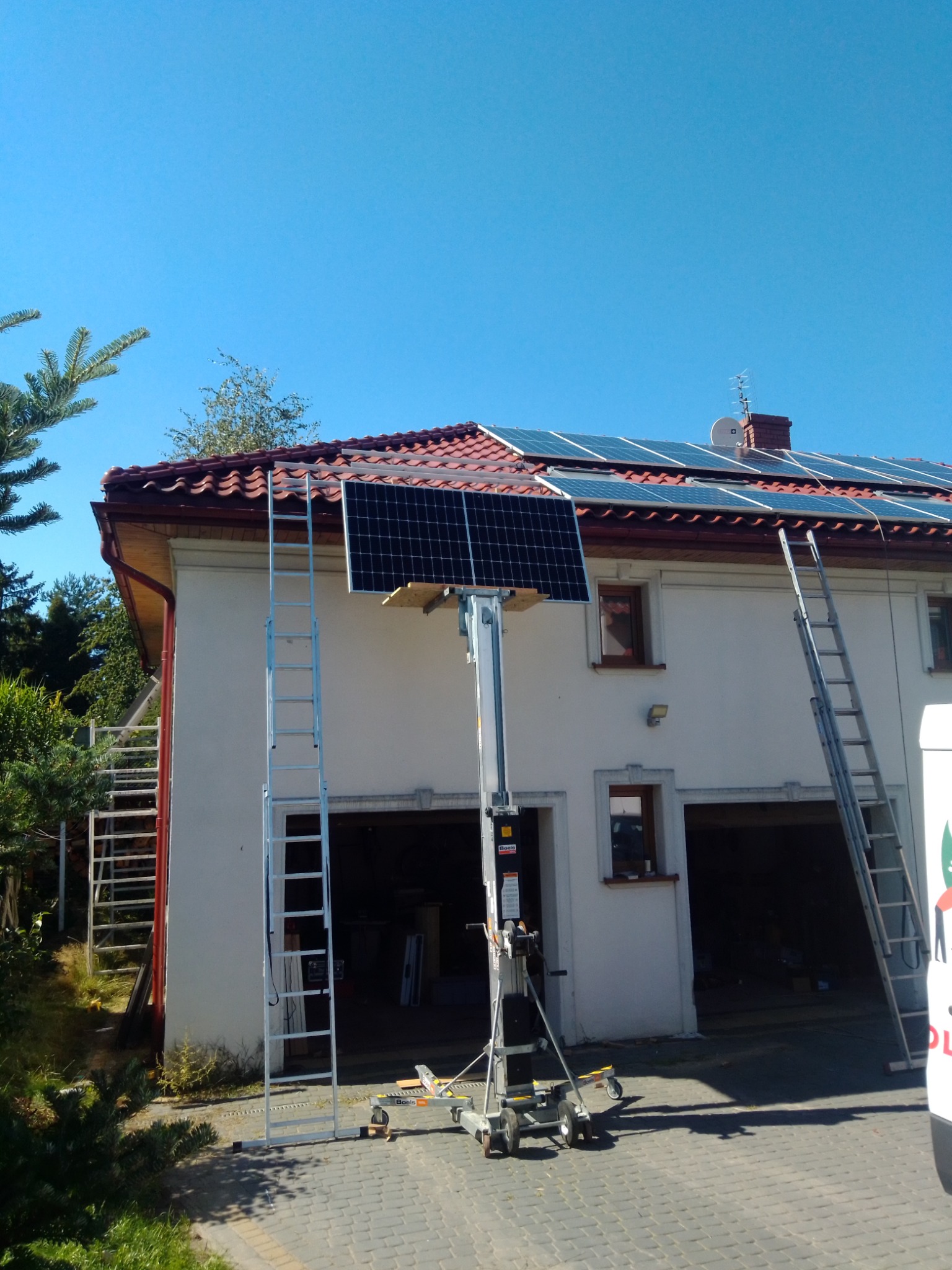 Montaż paneli na dachu dwupiętrowego domu z użyciem podnośnika i drabiny, błękitne niebo w tle.