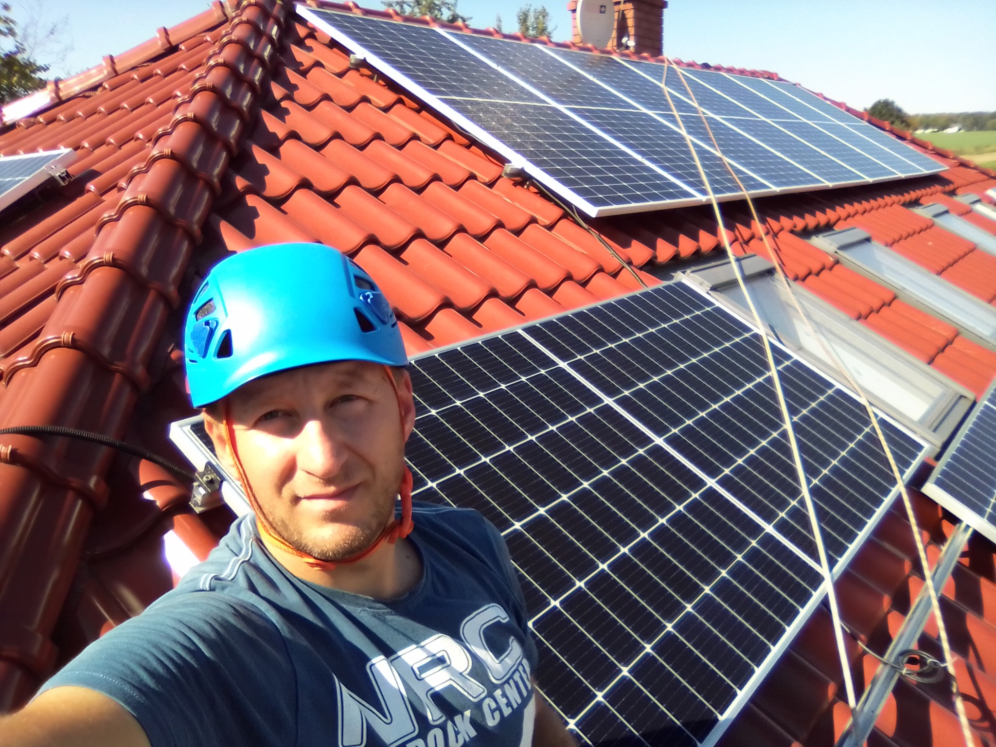 Mężczyzna w niebieskim kasku robi selfie na dachu z panelami słonecznymi na tle czerwonej dachówki i błękitnego nieba.