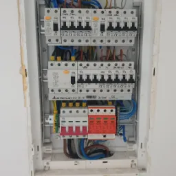 Wnętrze rozdzielnicy elektrycznej z zainstalowanymi zabezpieczeniami nadprądowymi, bezpiecznikami i ogranicznikami przepięć, widoczne kolorowe przewody i oznaczenia na urządzeniach.