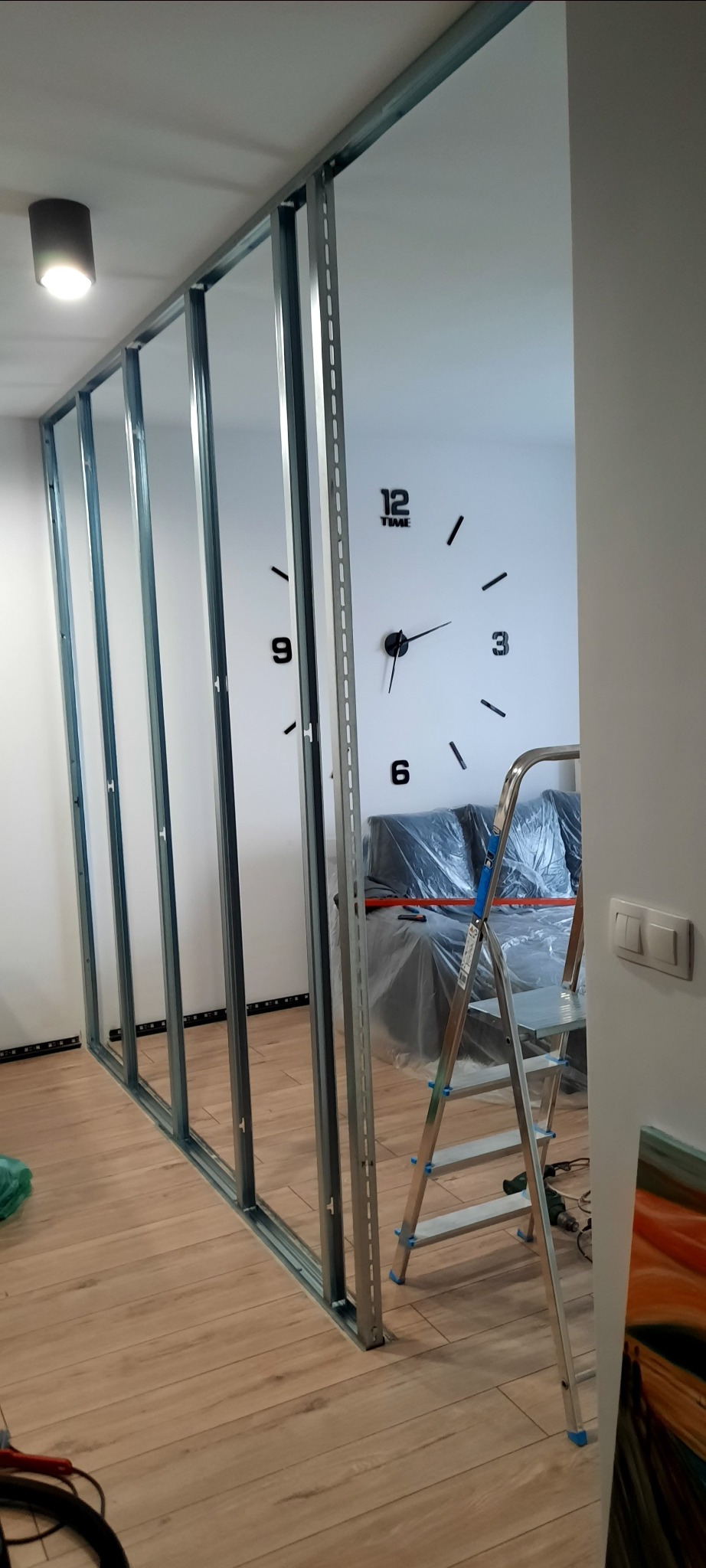 Wnętrze w trakcie montażu stelaża z profili stalowych do ścianki działowej, widoczna drabina aluminiowa, zabezpieczona folią kanapa i dekoracyjny zegar ścienny.