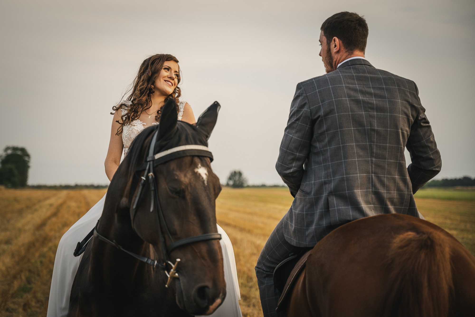 Panna młoda w białej sukni i pan młody w szarym garniturze siedzą na koniach na tle pola po żniwach, panna młoda uśmiecha się patrząc na pana młodego.