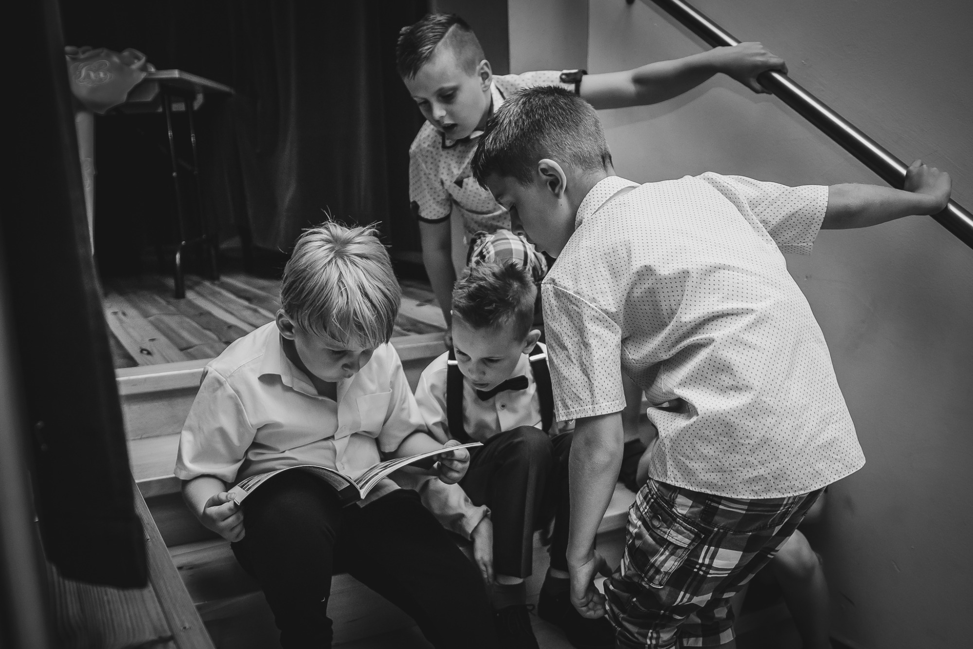 Czarno-białe ujęcie grupy chłopców na schodach, jeden czyta książkę, pozostali spoglądają na nią z góry, jeden opiera się o poręcz.