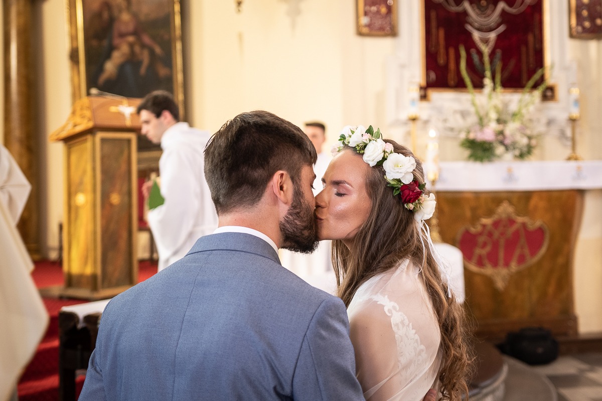 Pocałunek nowożeńców w kościele, panna młoda w wianku z białych i czerwonych kwiatów, jasna suknia z koronkowymi plecami, pan młody w błękitnym garniturze, w tle ołtarz i ksiądz.