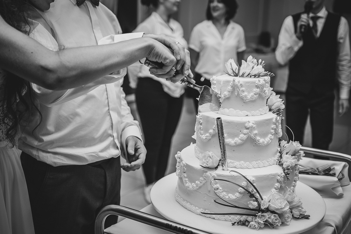 Czarno-białe ujęcie pary krojącej trzypiętrowy tort weselny, zdobiony kremem i kwiatami, z gośćmi i konferansjerem w tle.