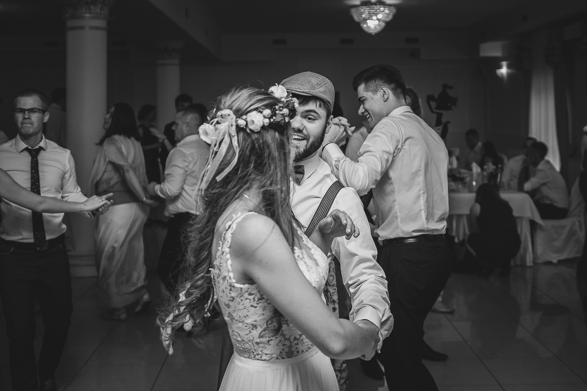Czarno-białe zdjęcie pary młodej tańczącej podczas wesela; panna młoda z wiankiem na głowie, pan młody w kaszkiecie i szelkach, w tle tańczący goście.