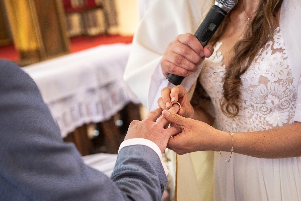 Moment nakładania obrączki podczas ceremonii ślubnej, w tle biała suknia z koronkowymi zdobieniami i mikrofon w dłoni celebransa.