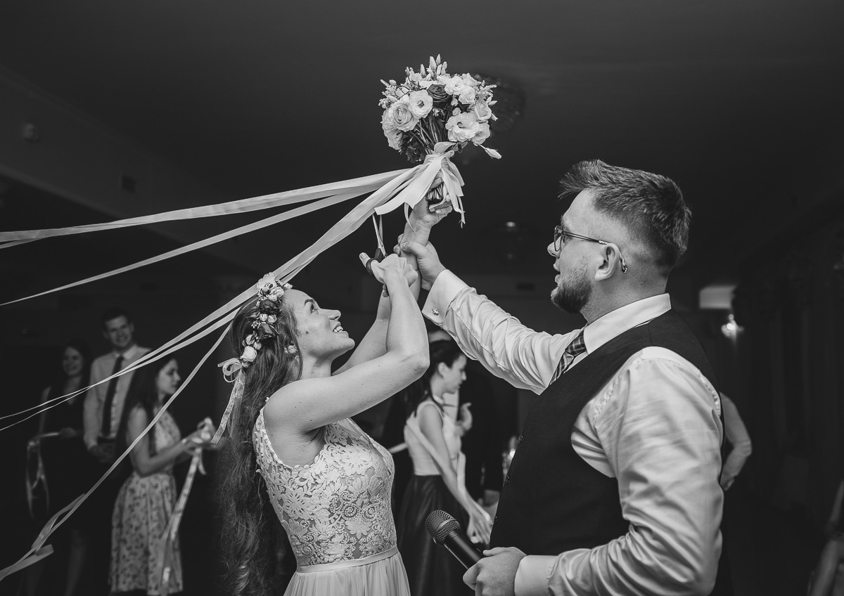 Czarno-białe zdjęcie panny młodej w koronkowej sukni i wianku, trzymającej bukiet z długimi wstążkami nad głową, z pomocą pana młodego w okularach i kamizelce, podczas tradycyjnego rzucania...