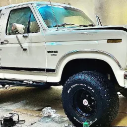 Biały Ford Bronco z podniesionym zawieszeniem i oponami terenowymi, widoczny w garażu na kartonach, podczas prawdopodobnych prac detailingowych lub naprawczych.