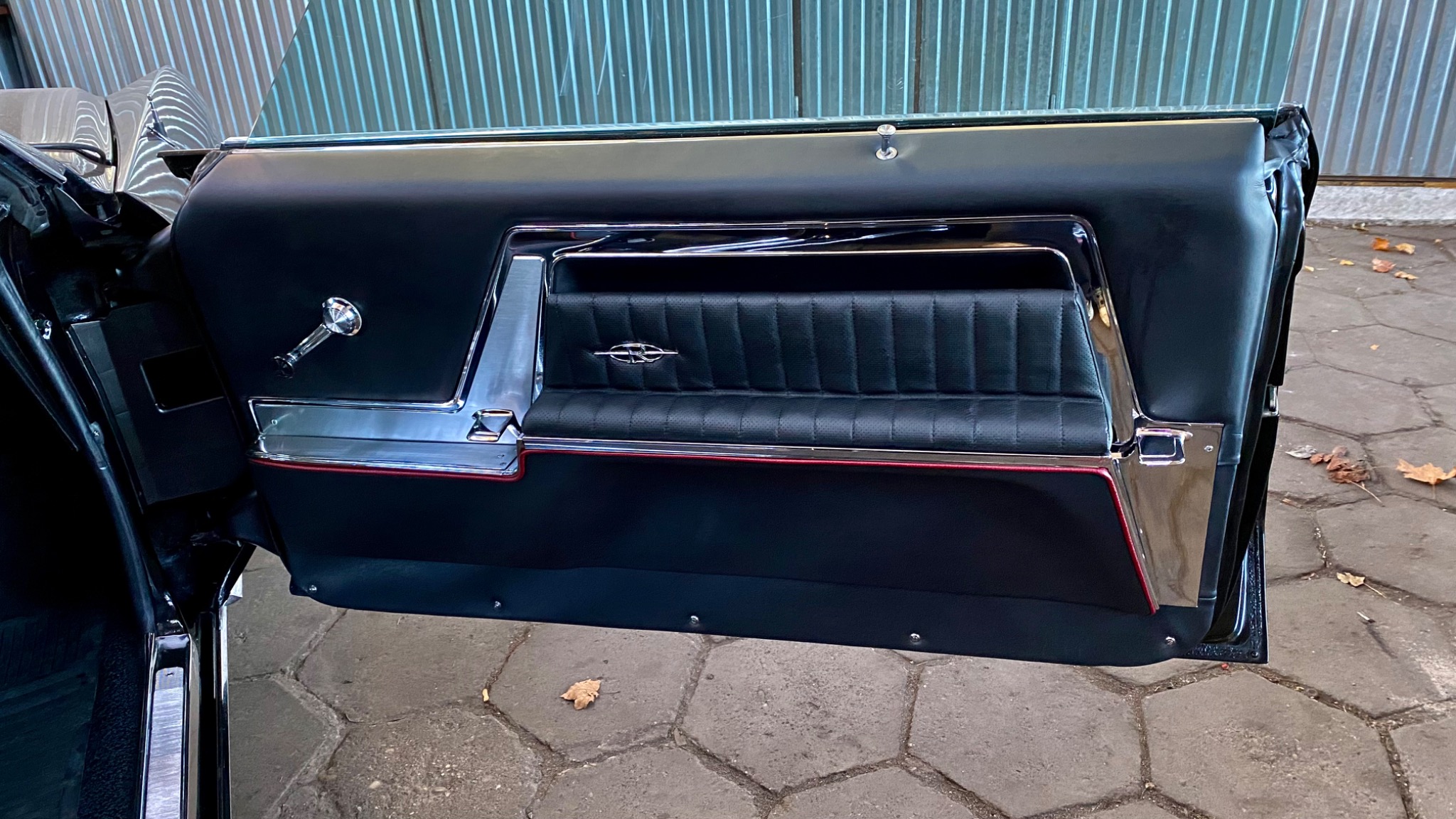 Wnętrze drzwi klasycznego samochodu z czarną tapicerką, pikowanym panelem i chromowanymi detalami, widok na klamkę i emblemat marki.