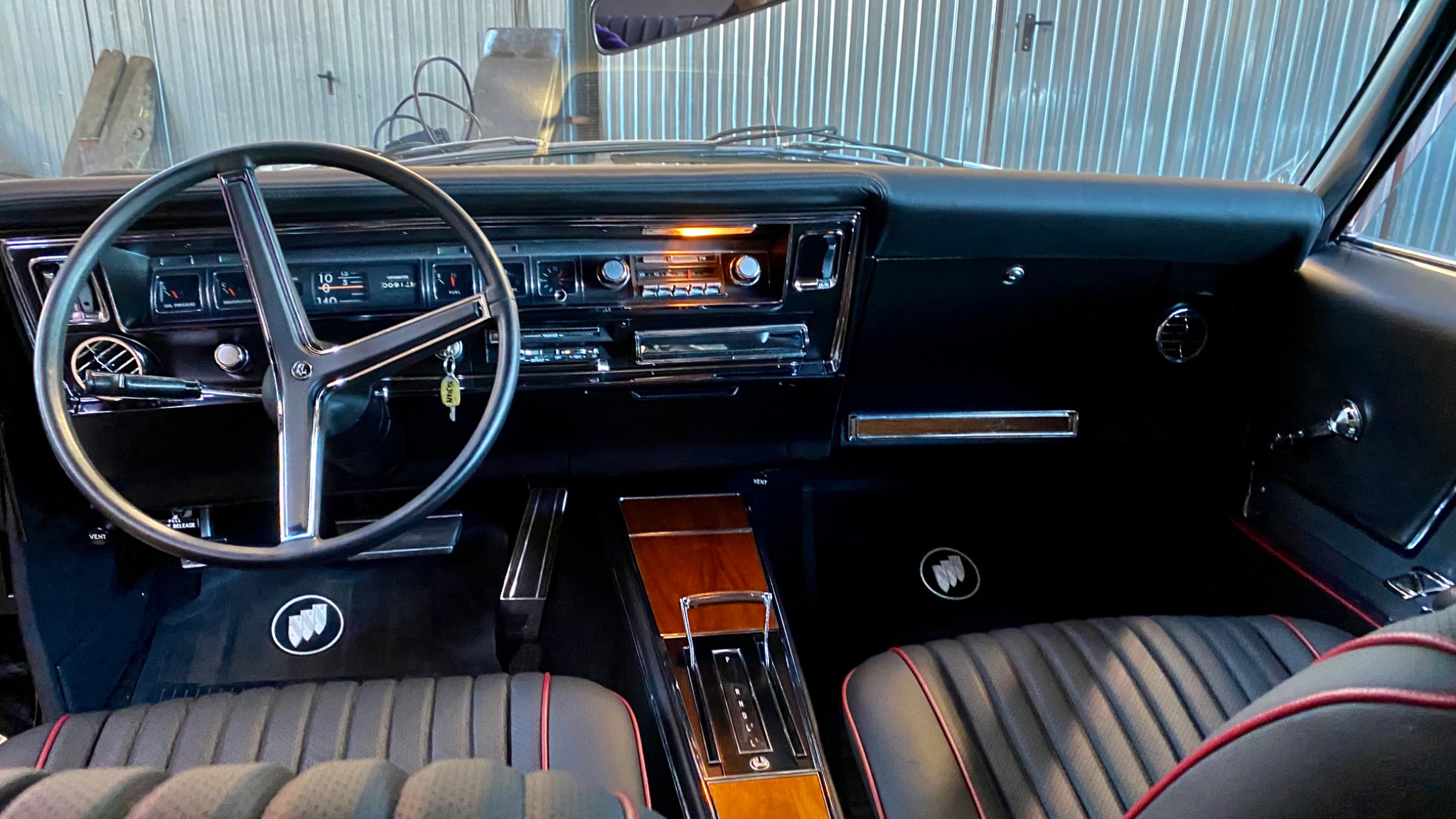 Wnętrze odrestaurowanego klasycznego samochodu Buick Riviera z czarną tapicerką z czerwonymi akcentami, chromowaną kierownicą i deską rozdzielczą z włączonym radiem.