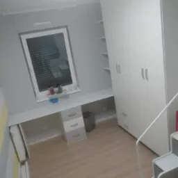 Pokój dziecięcy z białymi meblami: szafa trzydrzwiowa z półkami, biurko z szufladami i grzejnikiem pod oknem, widok z antresoli.