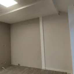 Wnętrze biura po malowaniu ścian na beżowo, z widocznymi fragmentami podłogi z paneli i oknem z naklejką. Świeżo pomalowane ściany, przygotowane do dalszych prac wykończeniowych.