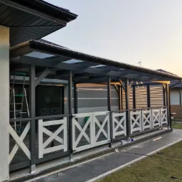 Zadaszona drewniana weranda z białymi krzyżowymi balustradami i szarą konstrukcją nośną, widok z zewnątrz w trakcie zachodu słońca, drabina oparta o ścianę budynku.