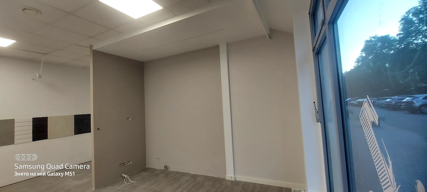 Wnętrze biura po malowaniu ścian na beżowo, z widocznymi fragmentami podłogi z paneli i oknem z naklejką. Świeżo pomalowane ściany, przygotowane do dalszych prac wykończeniowych.